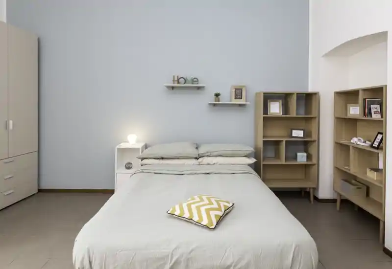Camera da letto luminosa e minimale con letto matrimoniale al centro, pareti dai toni pastello e scaffalature in legno. L'ambiente è tranquillo e ben curato, perfetto per annunci.