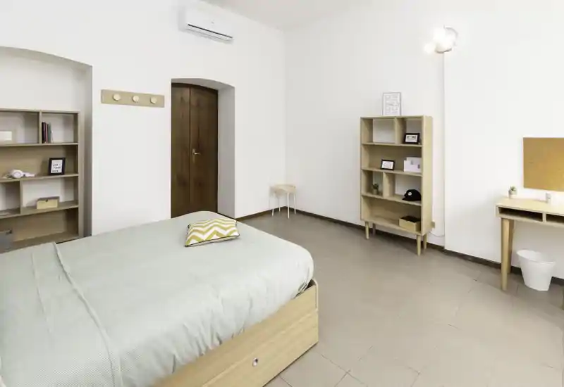 Camera da letto luminosa e minimal con un letto matrimoniale ben rifatto, mobili in legno chiaro e biancheria verde tenue. Ambiente arioso e ordinato con scaffali e una piccola scrivania.