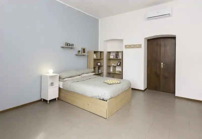 Camera da letto luminosa e minimal con letto in legno completo, biancheria verde chiaro, comodino con lampada e mensole semplici; parete pastello e porta in legno creano un'atmosfera tranquilla perfetta per gli annunci.