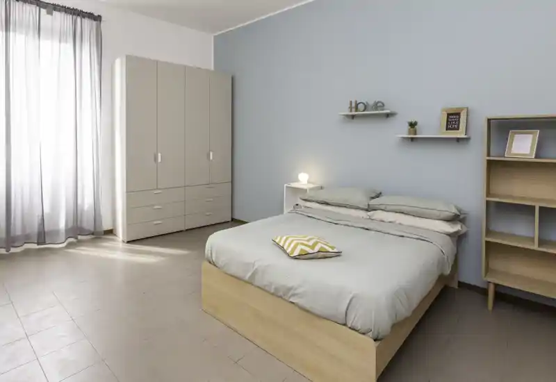 Camera da letto moderna e luminosa con letto matrimoniale ben rifatto, parete azzurro pastello, armadio e mensole; la luce naturale crea un ambiente tranquillo e arioso.