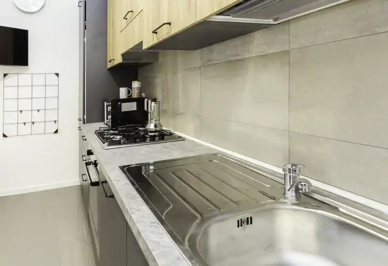Cucina moderna a corridoio con lavello in acciaio inox, piano cottura a gas e pensili in legno chiaro; piano pulito e rivestimento neutro adatto a piccoli appartamenti.