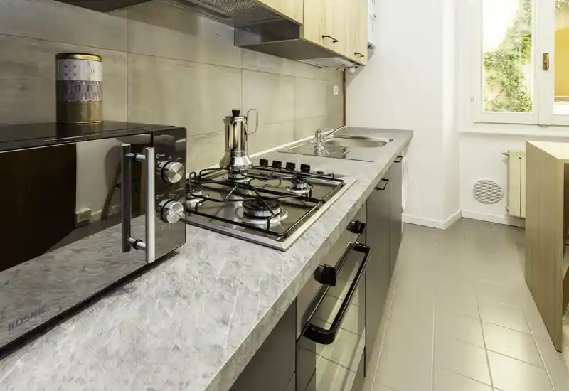 Cucina moderna a corridoio, luminosa, con piano cottura a gas, forno incassato, microonde e lavello in acciaio; mobili in legno chiaro e finestra che garantisce luce naturale.