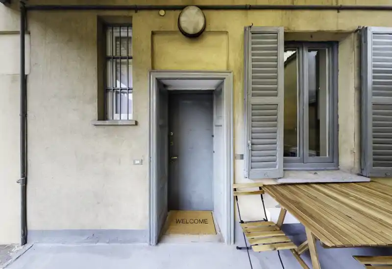Accogliente ingresso con zerbino "WELCOME", porta grigio-blu e persiane; un piccolo tavolo e sedie a destra suggeriscono un'area esterna accogliente.