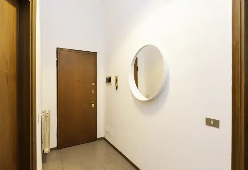 Ingresso luminoso e semplice con porta principale, specchio rotondo a parete e pavimento piastrellato — un atrio ordinato e accogliente.