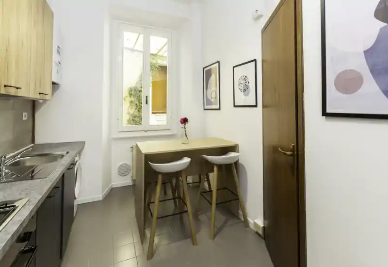 Cucina moderna compatta con piccolo bancone in legno, due sgabelli, lavello e elettrodomestici integrati. Finestra luminosa e accenti decorativi essenziali creano un ambiente accogliente.