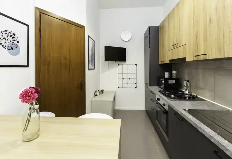 Cucina compatta e moderna con pensili in legno, basi scure, forno integrato e piano cottura a gas, più un piccolo tavolo con vaso di fiori — luminosa, ordinata e ben allestita per annunci.