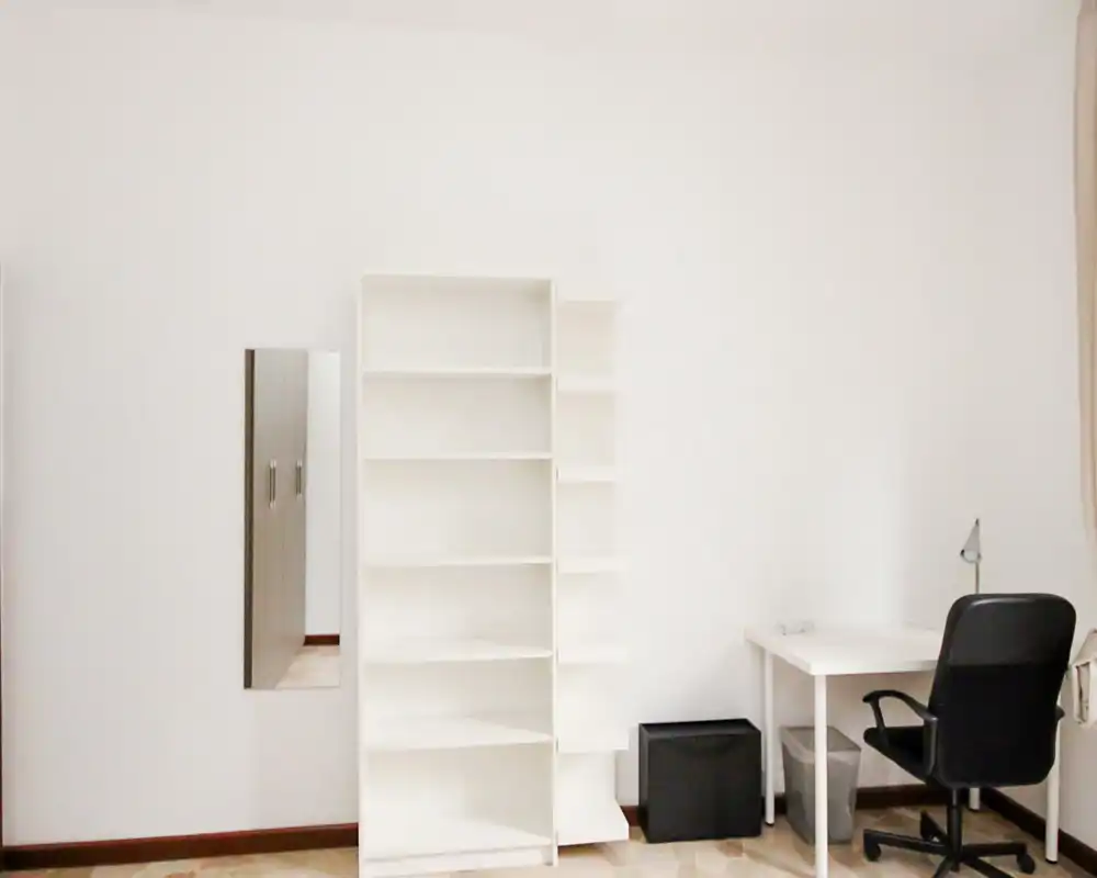 Zona de estar luminosa y minimalista con estanterías blancas, escritorio y silla de oficina: un espacio versátil para trabajar o descansar.