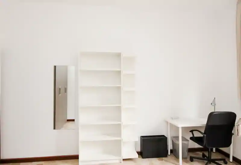 Zona de estar luminosa y minimalista con estanterías blancas, escritorio y silla de oficina: un espacio versátil para trabajar o descansar.