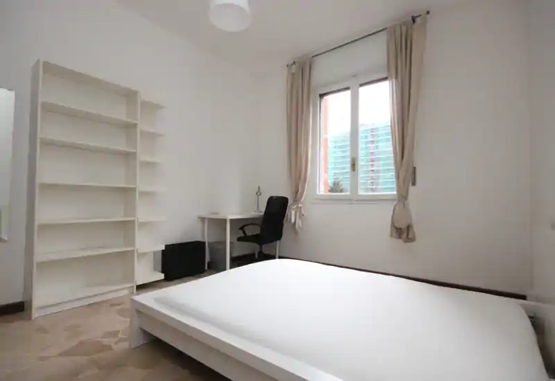 Dormitorio luminoso y minimalista con una gran cama baja, estantería y un pequeño escritorio junto a la ventana — decoración blanca y luz natural.
