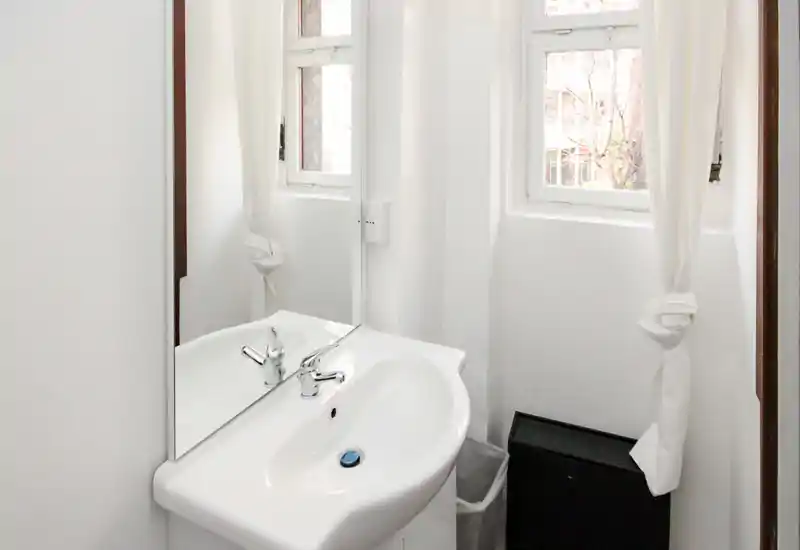 Bagno compatto bianco con lavabo a parete, rubinetto cromato e specchio; due piccole finestre con tende annodate che lasciano entrare luce naturale.