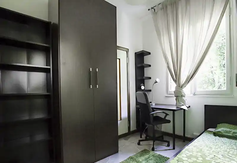 Camera da letto compatta e luminosa con letto singolo, armadio scuro e una scrivania vicino alla finestra—linee pulite e accenti verdi creano un angolo tranquillo.