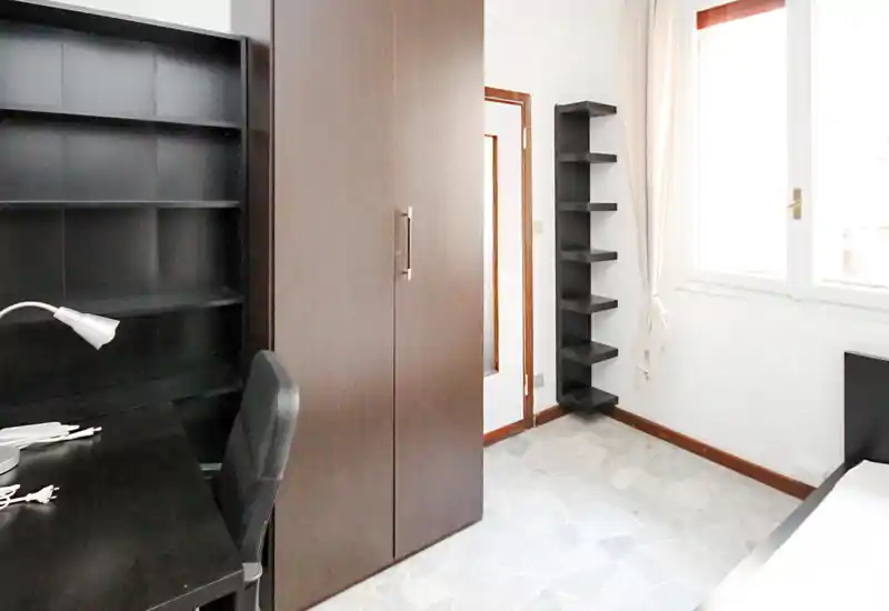 Piccola camera da letto con mobili in legno scuro: armadio, scrivania e vista parziale del letto vicino alla finestra. Luce naturale abbondante e layout semplice e funzionale, ideale per una persona.