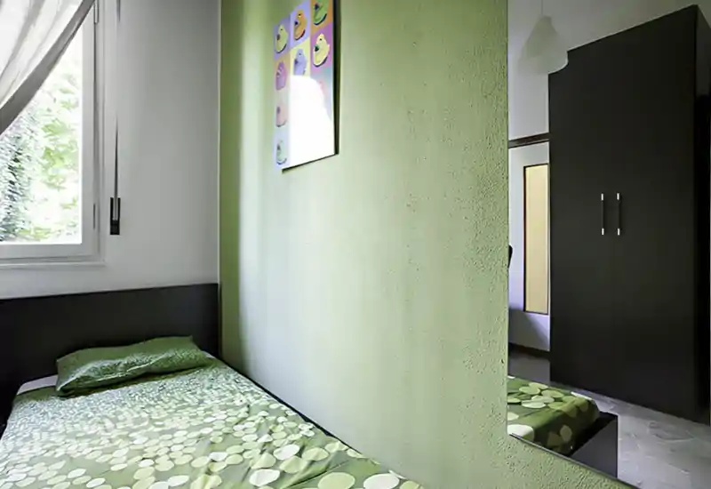 Camera singola compatta con parete verde, biancheria a fantasia verde e finestra che illumina naturalmente l'ambiente. Testiera scura e armadio visibile nello specchio, soluzione ordinata e accogliente per una persona.