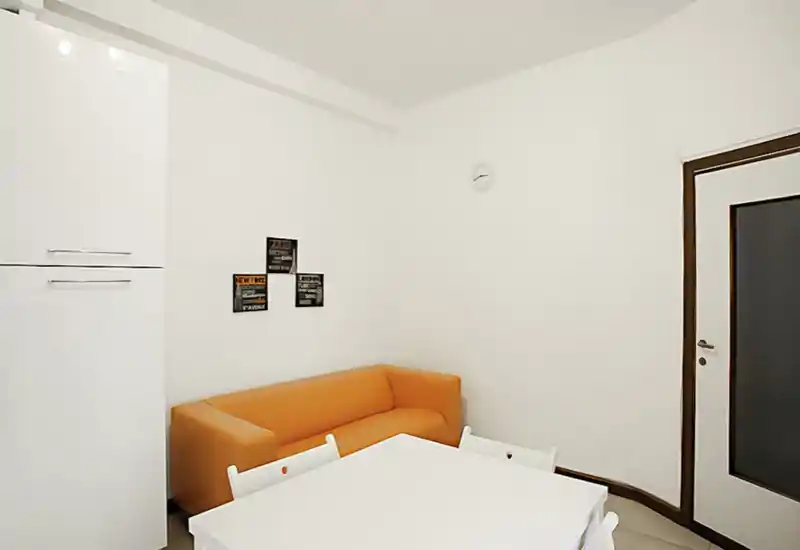 Area living compatta e luminosa con divano arancione moderno, piccolo tavolo da pranzo e arredi bianchi minimal — ideale per un monolocale accogliente.