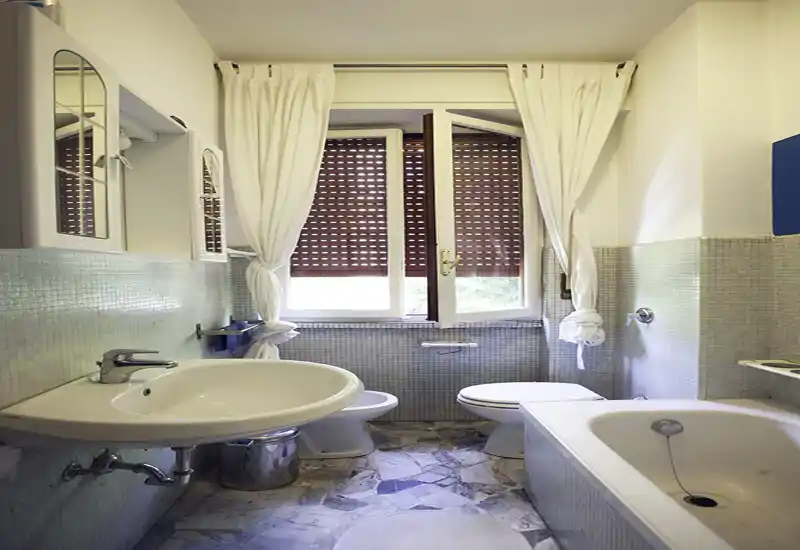 Bright, well-lit bathroom with a wall-mounted sink, bathtub and toilet, pale mosaic tiles and a large window with curtains letting in natural light.