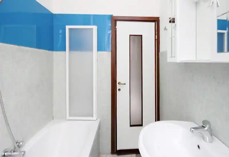 Clean, compact bathroom with a bathtub, handheld shower, sink and a white door—bright, tiled walls and simple modern fixtures.