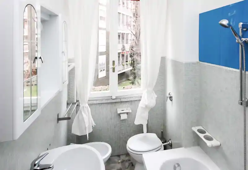 Compact, bright bathroom with mosaic tile walls, a shower head, toilet and bidet visible; a large window with sheer curtains provides natural light.