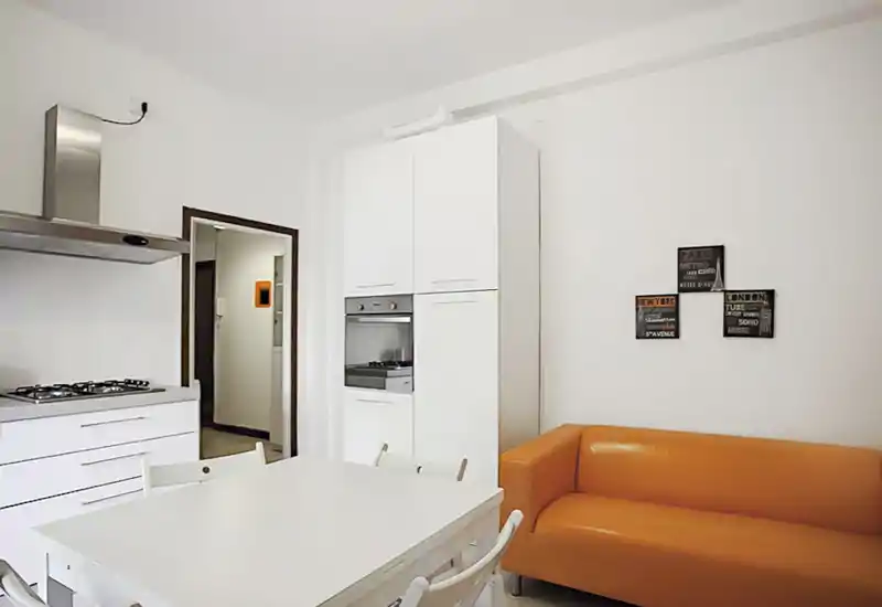 Bright modern open-plan kitchen with white cabinetry, built-in oven and a small dining table; an orange sofa adds a warm accent and contemporary feel.