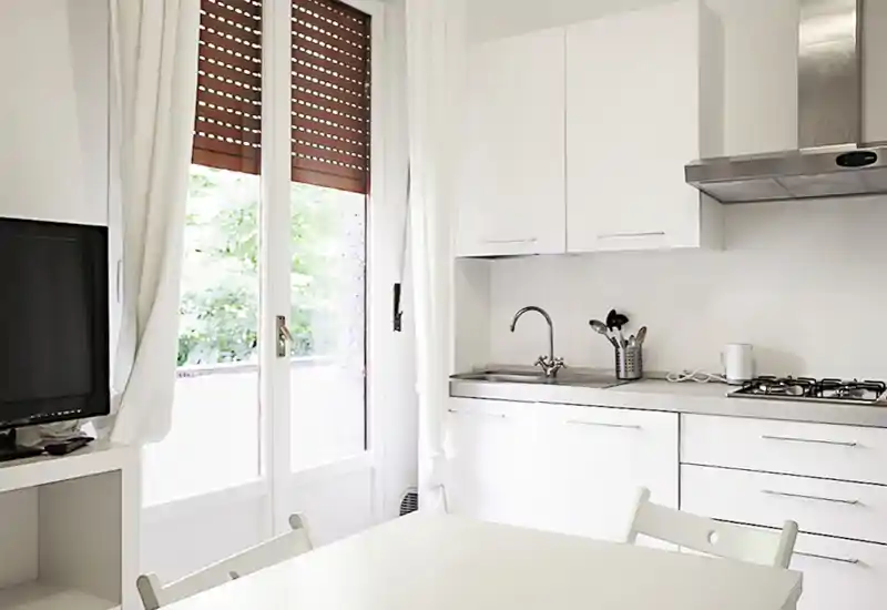 Bright, modern kitchen corner with white cabinets, stainless sink and gas hob, plus a small dining table by a large glass door letting in natural light.