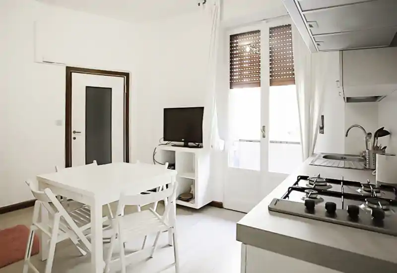 Angolo cottura luminoso e compatto con piano cottura a gas, lavello e un piccolo tavolo da pranzo per quattro vicino alla porta balcone — ideale per un appartamento cittadino accogliente.