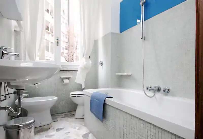 Bright, clean bathroom with a bathtub, wall-mounted showerhead, sink and toilet; white mosaic tiles and a window provide natural light.