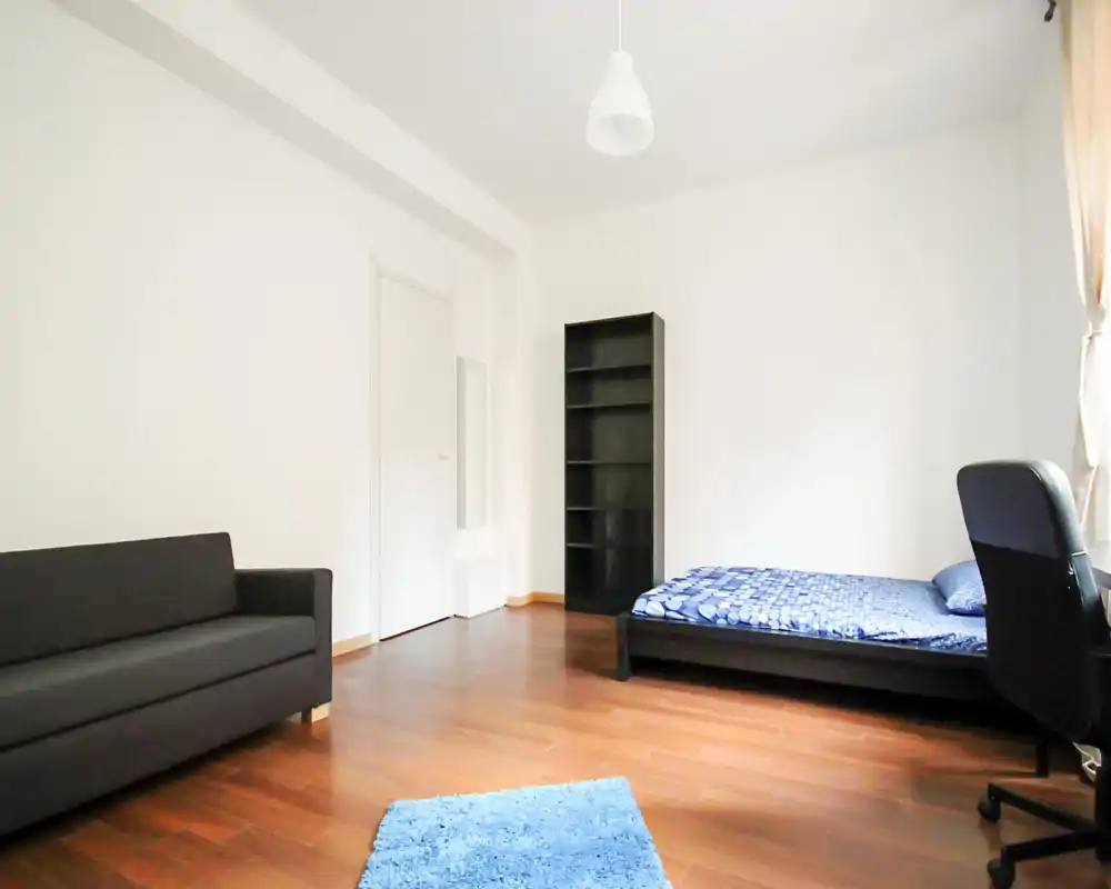 Dormitorio luminoso y minimalista con cama baja, sofá, estantería y suelos de madera. Paredes blancas y luz natural que ofrecen una sensación limpia y moderna.