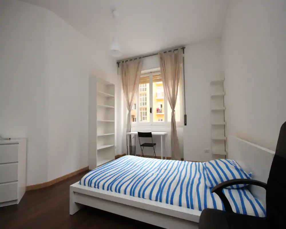 Camera da letto luminosa e minimal con letto matrimoniale, mobili bianchi e una scrivania davanti alla finestra; linee pulite e luce naturale ideali per gli annunci.