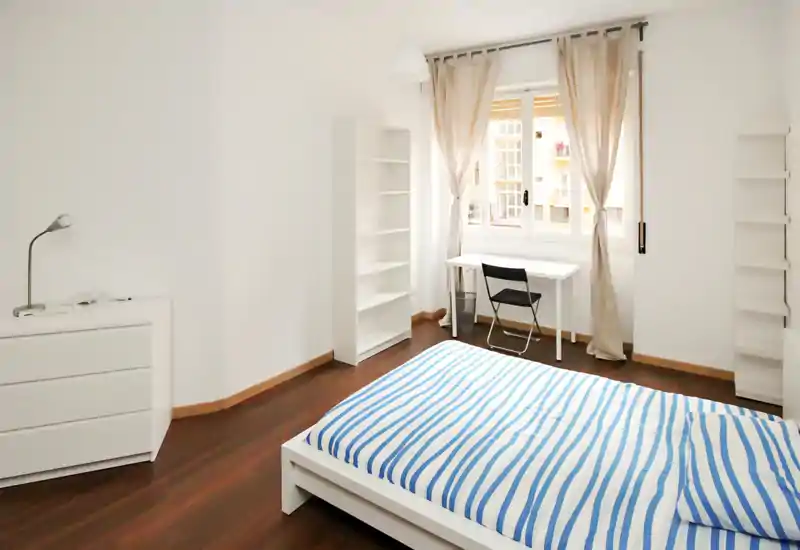 Camera da letto luminosa e minimal con letto basso bianco, biancheria a righe, scrivania vicino alla finestra e scaffalatura — perfetta per un annuncio di affitto.