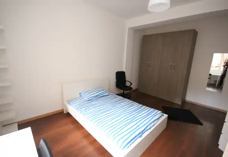 Camera da letto luminosa e minimal con letto singolo, biancheria a righe, armadio e piccola area scrivania — pulita e funzionale per annunci.