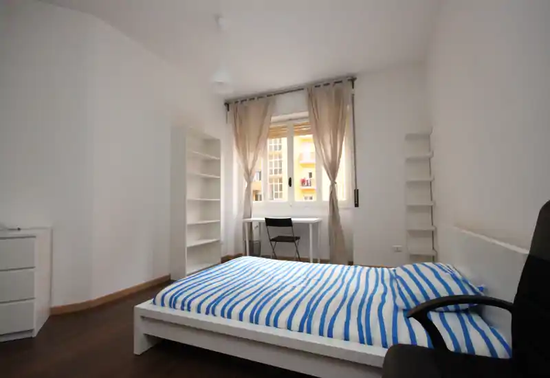 Camera da letto luminosa e minimal con letto matrimoniale, mobili bianchi e una scrivania davanti alla finestra; linee pulite e luce naturale ideali per gli annunci.