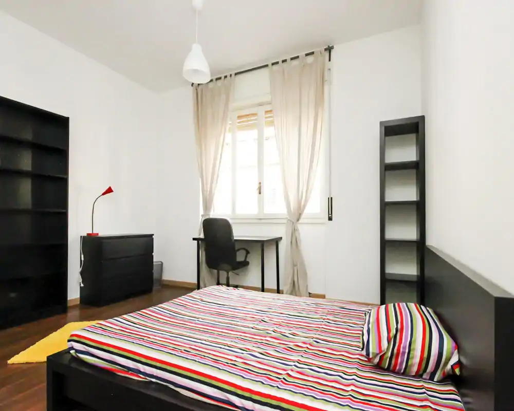 Bright, modern bedroom with a low black bedframe and colorful striped bedding, a desk by the window and minimal shelving — ideal for a single occupant or student.