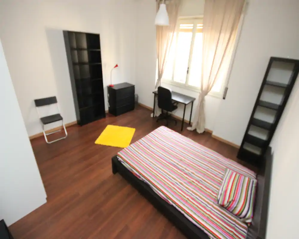 Bright, modern bedroom with a low platform bed dressed in colorful striped bedding, a desk by the window and open shelving — ideal for a student or professional.