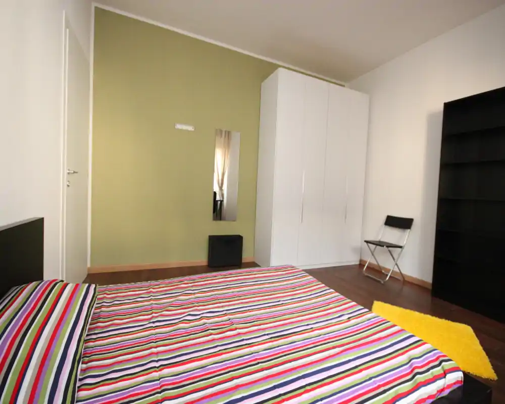 Bright, modern bedroom with a large striped bed, white wardrobe and minimalist furnishings — clean lines and colorful textiles give it a fresh, welcoming feel.