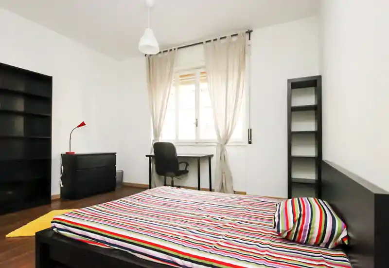 Bright, modern bedroom with a low black bedframe and colorful striped bedding, a desk by the window and minimal shelving — ideal for a single occupant or student.