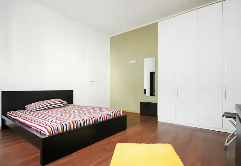 Bright, modern bedroom with a low-profile black bed, colorful striped bedding and a large white wardrobe. Clean lines, wooden floors and a small yellow rug create a simple yet inviting space.