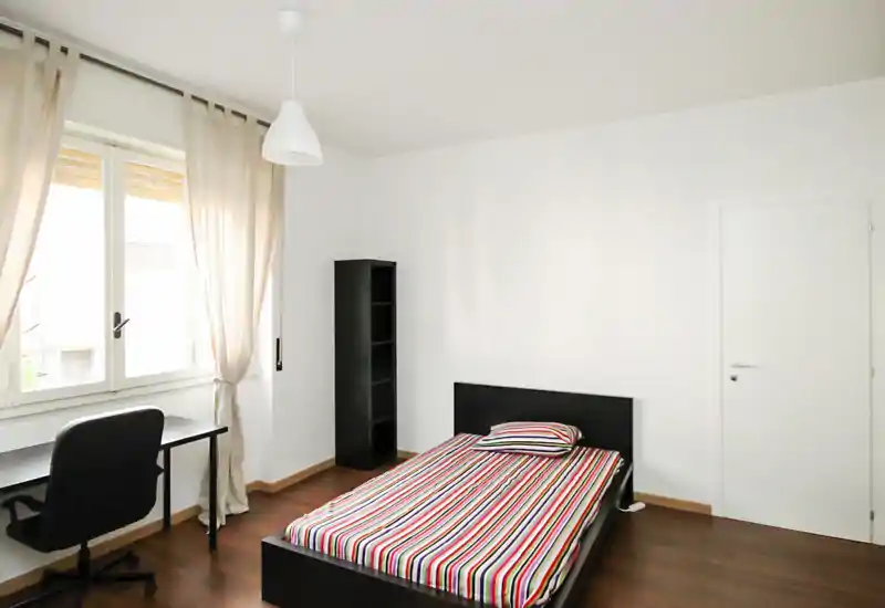Bright, minimal bedroom with a single bed, desk by the window and a tall shelving unit; clean white walls and natural light create an airy feel.