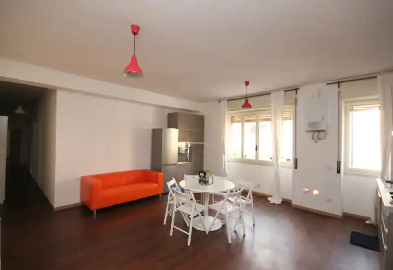 Espacio de estar luminoso y abierto con un sofá naranja pequeño, mesa redonda y sillas plegables bajo lámparas colgantes; grandes ventanas que aportan luz natural y un estilo moderno y minimalista.