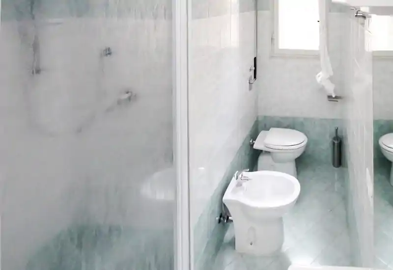 Cuarto de baño compacto y luminoso con bidé y inodoro visibles, suelos de azulejos turquesa claro y mampara de ducha opaca.