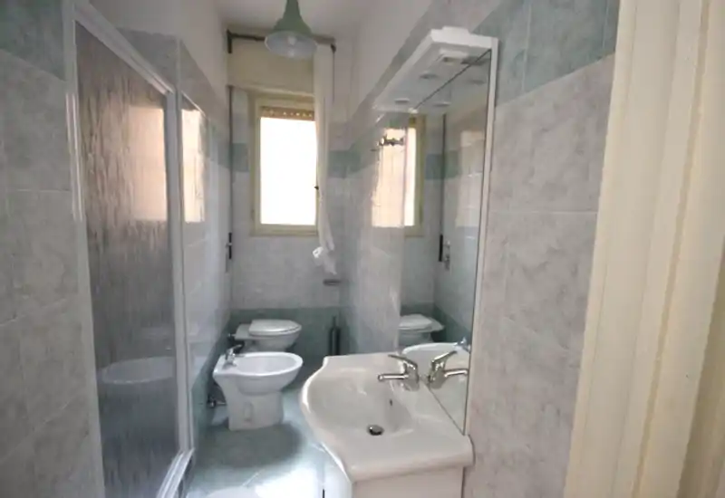 Baño estrecho completo con plato de ducha, bidé, inodoro y lavabo con espejo; paredes alicatadas y luz natural desde la ventana.