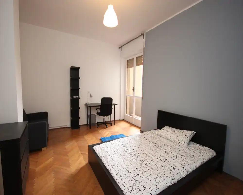 Camera da letto luminosa e minimal con letto matrimoniale, pavimento in parquet e una scrivania vicino alla finestra che affaccia su un piccolo balcone. Layout funzionale con arredi semplici e tonalità neutre.
