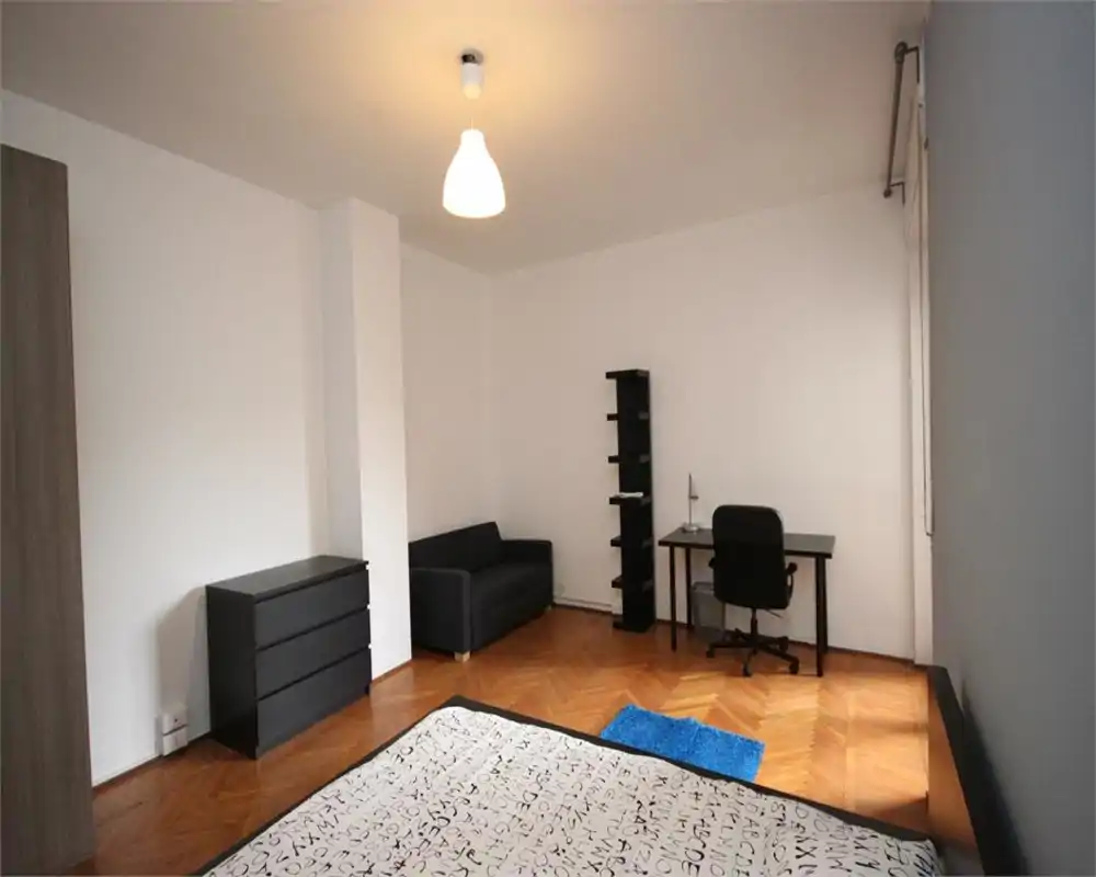 Camera da letto luminosa e minimal con letto matrimoniale in primo piano, piccolo divano, scrivania e scaffale: ideale per chi cerca uno spazio essenziale e funzionale.