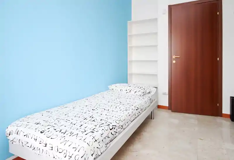 Habitación individual luminosa y minimalista con cama bien hecha frente a una pared azul claro y estanterías empotradas — estilo moderno y limpio, ideal para una persona.