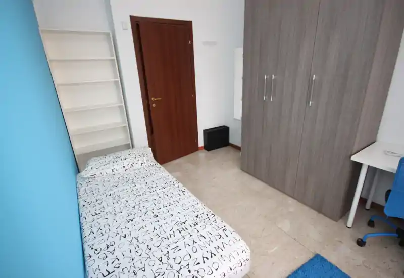 Dormitorio individual compacto con armario empotrado, pequeño escritorio y cama individual con ropa de cama estampada; espacio luminoso y ordenado, ideal para estudiante o persona sola.