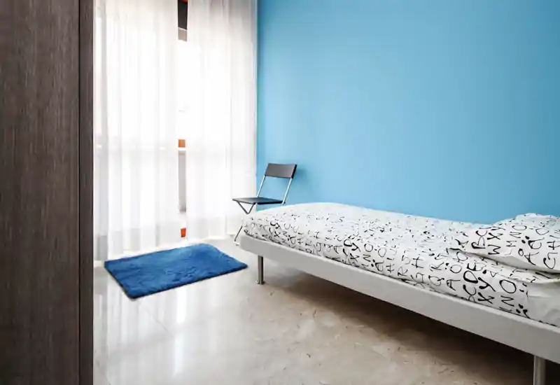 Dormitorio individual luminoso y minimalista con una cama blanca baja, ropa de cama con estampado y una pared azul. Cortinas traslúcidas dejan entrar luz natural suave y una pequeña alfombra aporta textura.