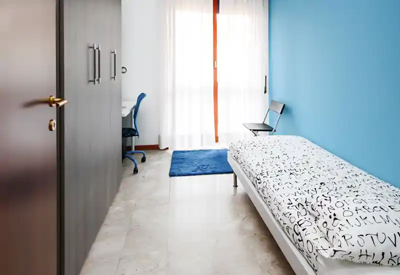 Dormitorio individual luminoso y minimalista con edredón estampado, pared azul y luz natural de una amplia ventana. Dispone de escritorio y silla, armario y una alfombra que aportan un aire moderno y acogedor.