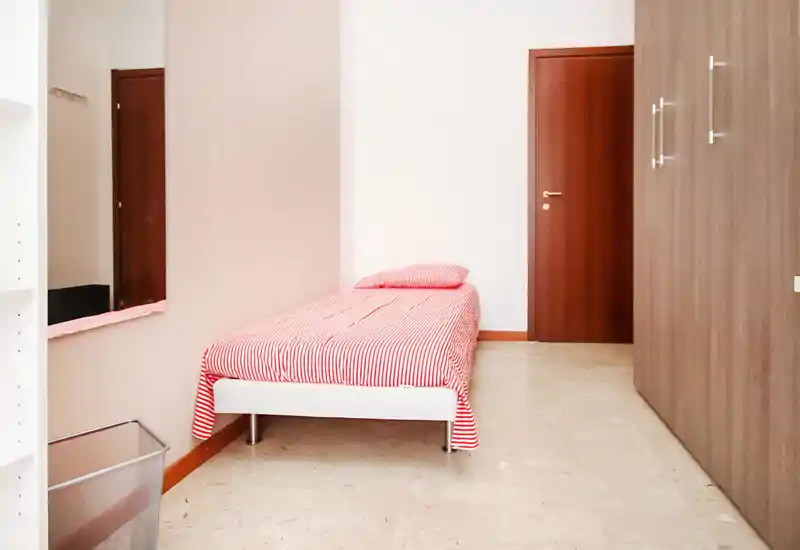 Habitación individual sencilla y ordenada con cama de rayas rojas, armario empotrado y paredes neutras — luminosa y sin desorden, ideal para una persona.