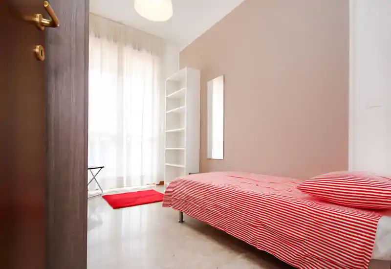 Dormitorio luminoso y minimalista con colcha de rayas rojas, gran ventana con visillos que aporta mucha luz natural y estantería sencilla.