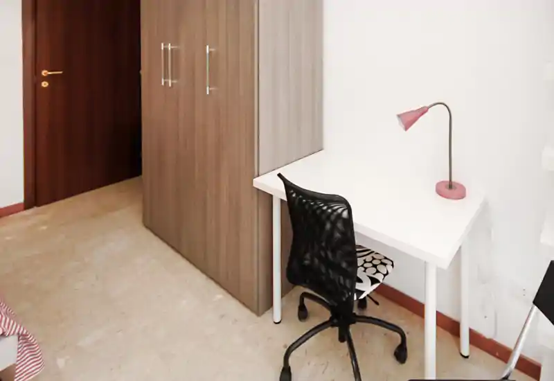 Rincón de dormitorio con escritorio blanco compacto, silla de malla negra y armario; decoración mínima y buena luz natural.