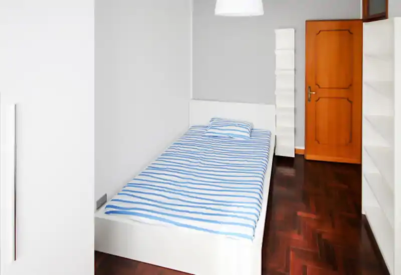 Dormitorio individual minimal y luminoso con cama blanca y ropa de cama a rayas azul y blanco, suelo de parquet y estanterías empotradas — luminoso y sencillo, perfecto para alquiler.