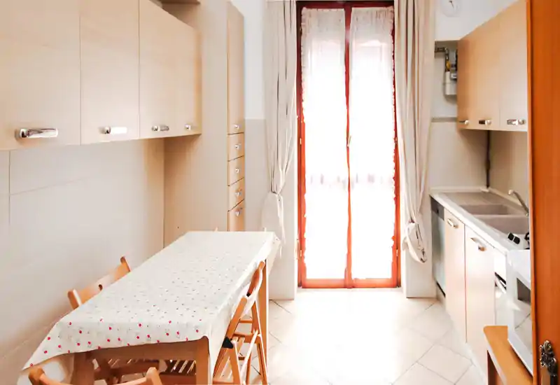 Cocina compacta y luminosa con armarios de madera, una pequeña mesa con mantel estampado y un fregadero junto a la puerta del balcón que aporta mucha luz natural.